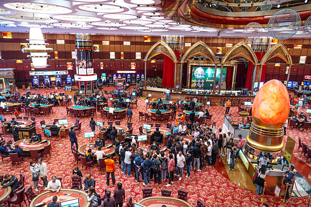 A bustling casino floor in Macau with numerous gaming tables, players, and dealers. Large stage backdrop, ornate arches, shimmering lights, and a glowing centerpiece create an opulent, entertainment-filled atmosphere.