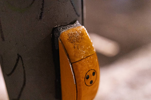 Close-up view of a yellow pedestrian crossing push button covered with frost and ice during freezing winter weather. The scene highlights hazardous icy conditions on public street infrastructure and the increased risk of slips and injuries during cold temperatures.