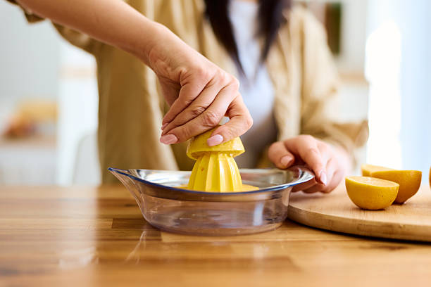 Woman's hands are manually squeezing a fresh lemon half with a yellow citrus reamer into a glass bowl on a wooden kitchen countertop, preparing a healthy drink or ingredient for cooking