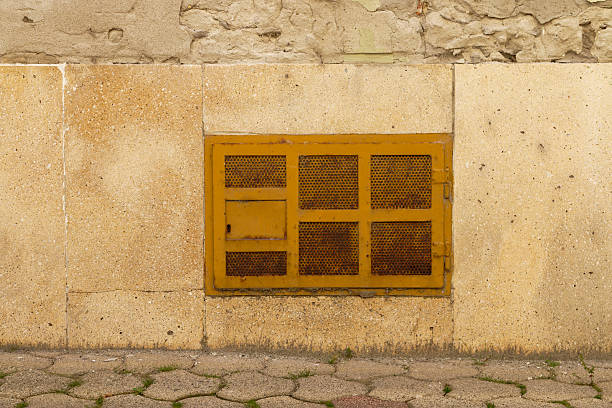 A close-up view of an old, rusty yellow metal grate embedded in a rough, beige stone wall, with a section of hexagonal cobblestone path in the foreground