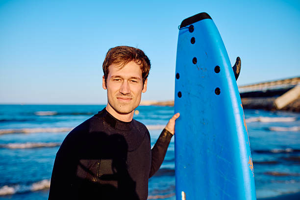 Young man in a wetsuit stands on the beach holding a blue surfboard, smiling confidently at the ocean before surfing session