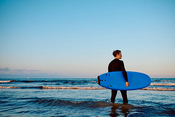 Young adult man in wetsuit holding a blue surfboard, looking out at waves during golden hour. Enjoying water sport