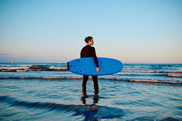 Young man in a wetsuit carrying a blue surfboard, looking out at the waves during a calm evening surf session