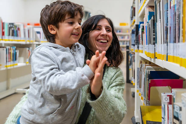 Mom and her happy child are smiling while looking at books on a shelf together, sharing a moment of learning and connection in a bright library environment