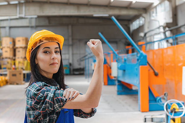 A confident young female factory worker wearing blue overalls and a yellow safety helmet flexes her arm with a smile inside a modern industrial manufacturing facility surrounded by heavy machinery metal processing equipment and industrial production lines symbolizing strength empowerment workplace safety and women in industry