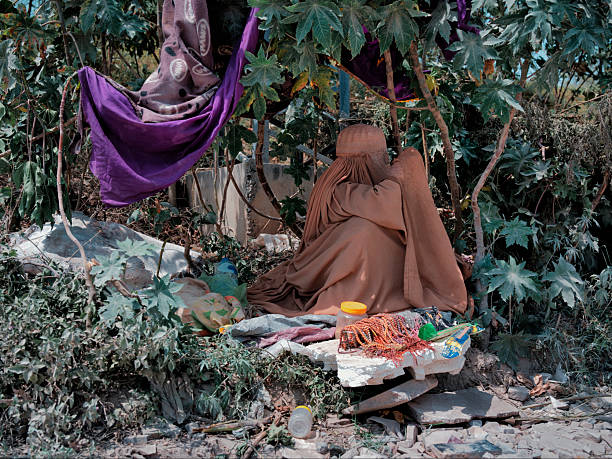 One woman in burqa sitting outdoors, finding shelter under foliage, representing vulnerability and hardship