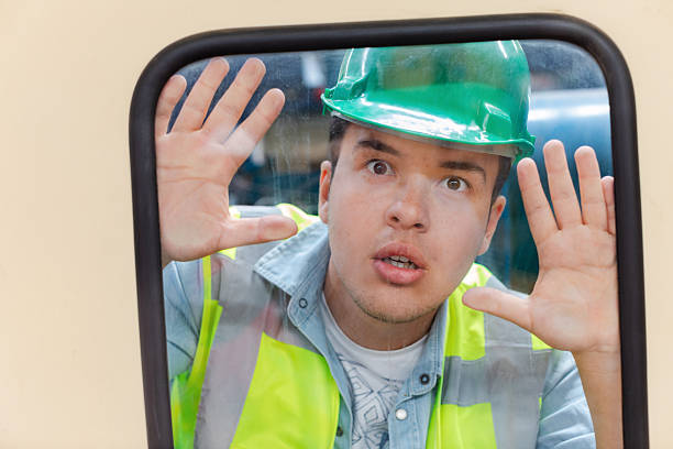 Young male industrial worker wearing green hard hat and high visibility safety vest pressing hands against a glass window with a shocked and curious facial expression inside an industrial workplace conveying humor surprise and safety awareness