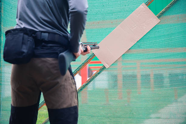 Adult male shooter aiming shotgun at cardboard target on a green mesh barrier