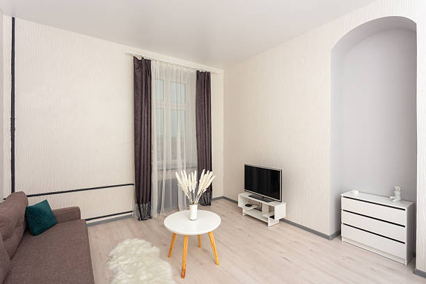 Bright minimalist living room with sofa, TV stand, round coffee table and window curtains. Modern apartment interior with light wood floor and clean white walls, ideal for rental listing.