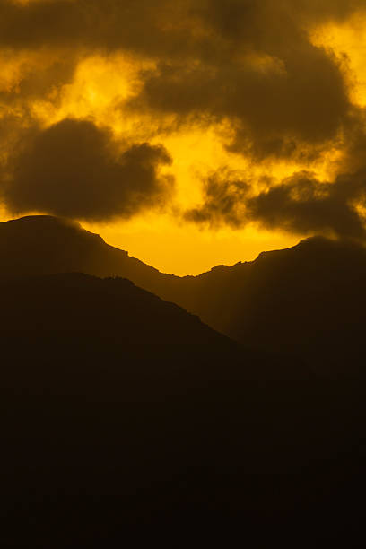 Glowing golden clouds illuminated by sunset above dark mountain slopes. Atmospheric evening scene with strong contrast.