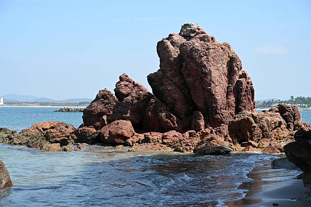 Prominent red rock formation emerging from shallow ocean water along the Mazatlán coastline. The image captures dramatic coastal geology, reflections on wet sand, and the rugged natural character of the Pacific shoreline.Keywords: rock formation, coastal rocks, shoreline, Mazatlán, Mexico, Pacific coast, seascape, natural landmark, geology, beach landscape, travel photography
