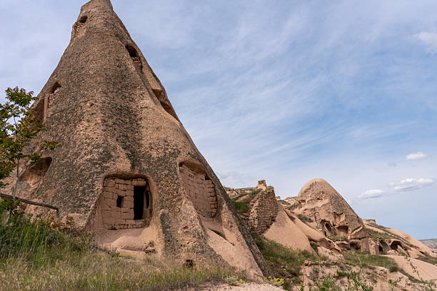 Tall conical fairy chimney with distinctive pointed cap rises prominently against sky in Pigeon Valley, Cappadocia, Turkey. The honey-colored volcanic tuff formation shows characteristic erosion with resistant darker cap protecting softer column beneath, with person visible at base providing scale reference. The dramatic formation represents Pigeon Valley's spectacular geological features, with this valley named for the extensive pigeon houses carved into rock faces where domesticated pigeons provided valuable fertilizer for agriculture in this semi-arid volcanic landscape.