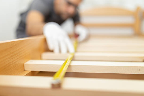 A person measures wooden slats for constructing a bed frame. The setting is a workshop filled with tools and materials. The focus is on accurate measurement.
