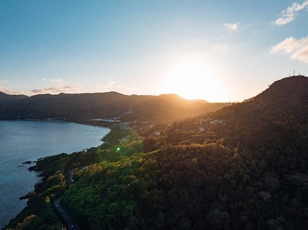 Golden hour sun setting behind lush green mountains, illuminating a winding coastal road and calm ocean waters around Mahe, Seychelles, creating a peaceful tropical evening scene