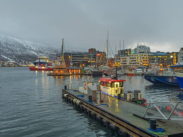 Tromso, Norway - December 23, 2025: The harbor and buildings along the boardwalk, from Stortorget. Plenty of small boats and mainland snow-covered mountain in back. SAR lifeboat in front