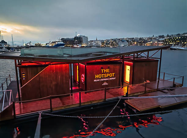 Tromso, Norway - December 23, 2025: The Hotspot floating sauna in the harbor, with slogan: relax, refresh, repeat. Island cityscape buildings in back, Red lights reflected in water
