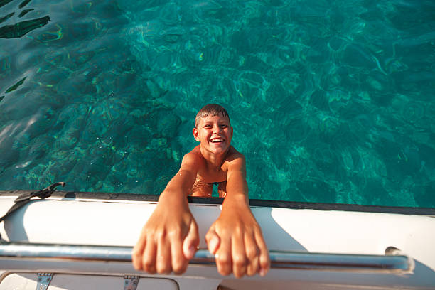Happy young boy smiling while holding the edge of a boat in crystal clear turquoise sea during summer family vacation in Greece.
