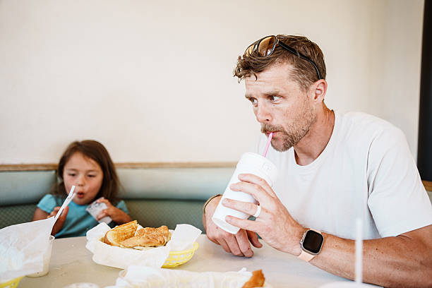 A father takes a sip of a milkshake while sitting next to his daughter in a retro-style diner booth.