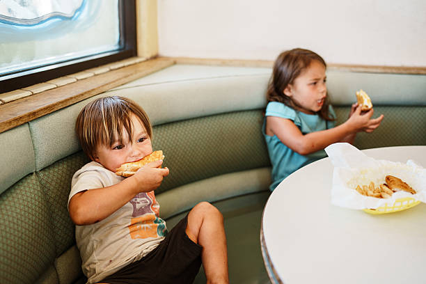 A two-year-old Eurasian boy and his older sister sit together in a diner booth and eat grilled cheese while stopping for lunch during a family road trip.