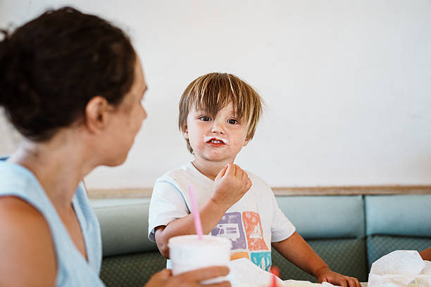 A two-year-old Eurasian boy with a milkshake mustache looks at the camera while sharing comfort food with his family at a small-town diner, during a summer road trip stop.