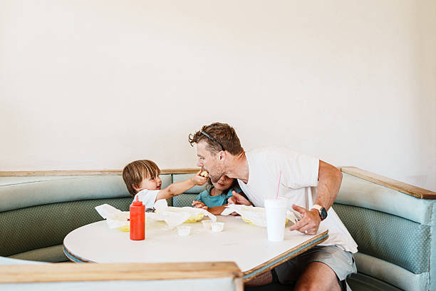 A Eurasian toddler boy reaches across a diner table to share a bite of his grilled cheese sandwich with his father, at a small-town retro diner during a summer family road trip.