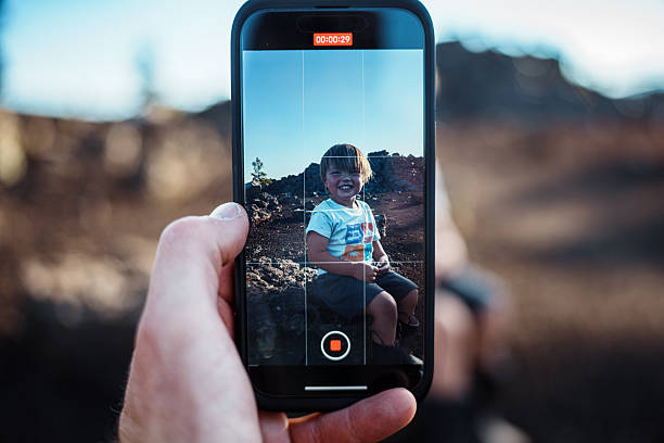The hand of an unrecognizable parent holds a smart phone which displays a cheerful toddler boy sitting on a rock, documenting a family hike in a volcanic landscape at Craters of the Moon National Monument.