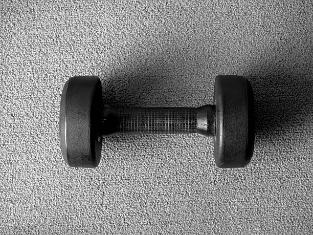 Single black dumbbell on grey carpet floor in minimal style. Concept of strength training, healthy lifestyle, weightlifting, fitness workout, and home gym discipline with focus on wellbeing.