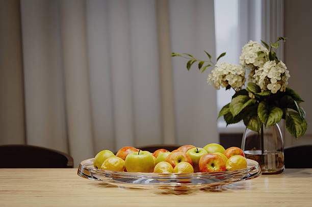 Digitally generated close-up of a glass bowl filled with apples and lemons on a wooden dining table, with a vase of white flowers and green leaves in soft focus behind. Warm indoor light and neutral curtains create a calm, contemporary atmosphere. Concept for healthy eating, hospitality, home cooking and cozy family life.