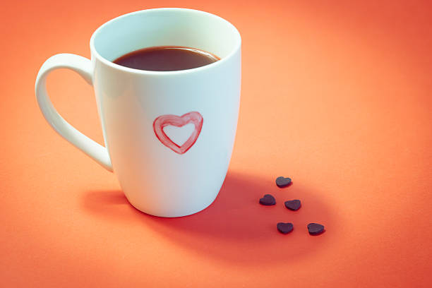 White ceramic coffee mug filled with coffee and featuring a red heart symbol on the front, placed on a bright orange background. Small heart-shaped confetti pieces are arranged beside the mug. Clean, minimal composition with copy space, suitable for Valentine’s Day themes, love and relationship concepts, romantic greeting cards, café promotion, and seasonal marketing materials.