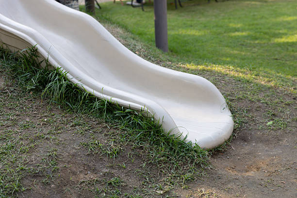 A white slide is on a grassy hill in public playground. The slide is covered in grass and dirt. The grass is green and the dirt is brown.