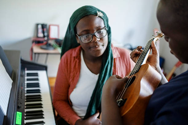 Sister wearing hijab teaching brother playing ukulele next to a piano