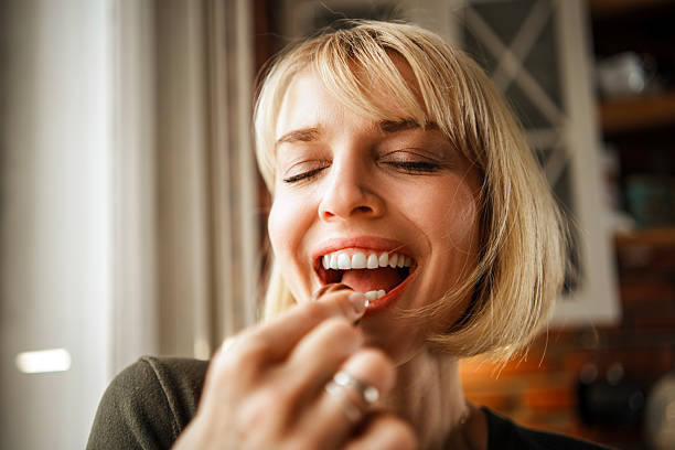 A cheerful blonde woman enjoys a chocolate praline while holding a plate of assorted chocolates. She is standing in a modern kitchen featuring white cabinetry and a rustic red brick wall.