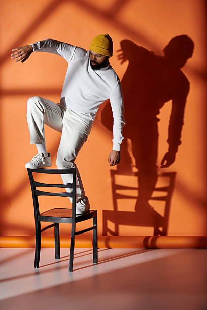 A fashionable young man playfully balances on a chair while striking a confident pose.