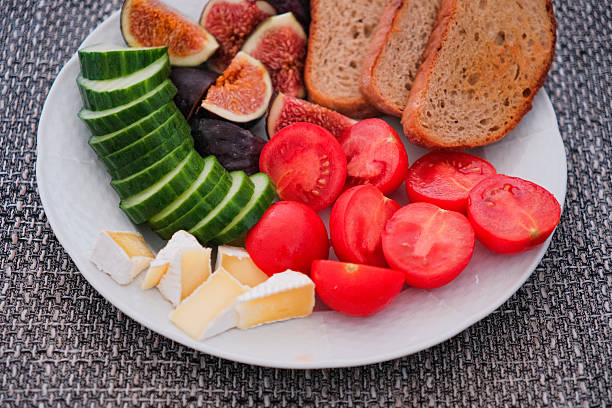 A nutritious and colorful breakfast platter featuring sliced fresh figs, cucumbers, cherry tomatoes, creamy brie cheese, and rustic bread, beautifully presented on a white plate in soft natural light against a textured grey background.