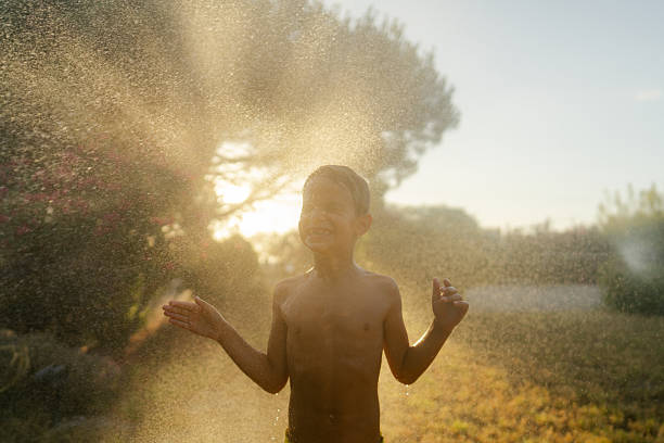 A front view shot of a young boy playing outdoors, he is having fun spraying water out of a water hose.
