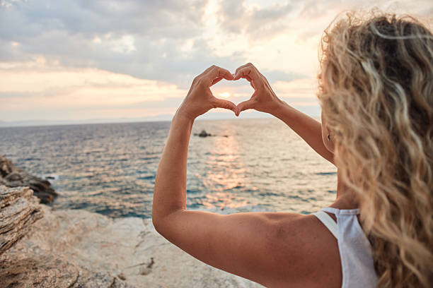 Woman making a heart sign with hands framing the sun at sunset over the ocean