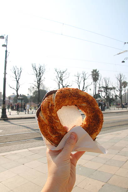 A POV shot of a single hand holding a traditional Turkish sesame bread ring (Simit) overflowing with rich chocolate hazelnut spread. The background features a clear, bright blue sky and the silhouette of iconic Ottoman-style street lamps. This image perfectly captures the indulgence of Turkish street food and the sunny atmosphere of a day out in Istanbul.