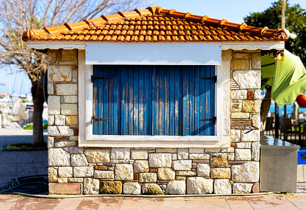 Mediterranean style stone buffet with a red tiled roof and blue wooden shutters, empty white banner mockup on the facade for cafe or boutique branding in a coastal area.
