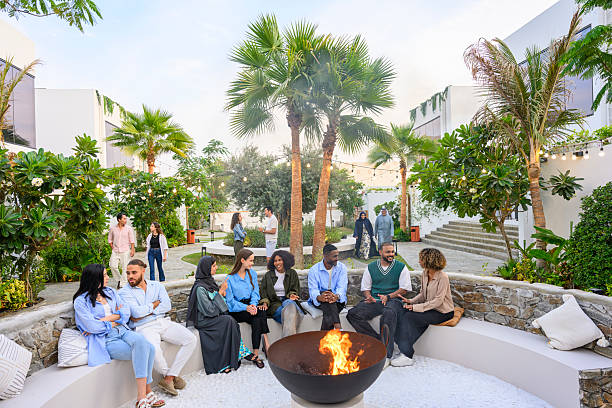 Group of diverse friends gathering around a warm bonfire, relaxing and engaging in conversation within a modern outdoor lounge while surrounded by lush greenery and palm trees