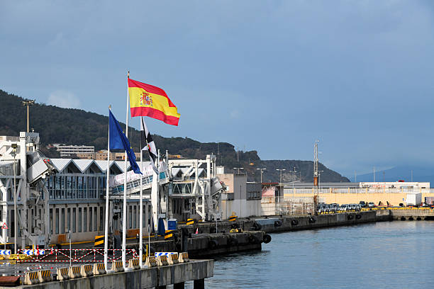 Ceuta, Spain: The national flag of Spain flies from a pole at the rhe Maritime Station (Estación Marítima) of the Port of Ceuta, the principal passenger fery terminal located on the Muelle Cañonero Dato. It serves as the critical transportation hub connecting the Spanish autonomous city in North Africa with the Port of Algeciras on the Iberian Peninsula. Handling nearly two million passengers annually, the facility is vital for regional connectivity and the logistical management of the \