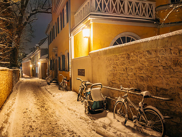 A quiet, snow-covered street in Weimar, Germany at night, illuminated by warm lamps. Bicycles line a stone wall beside a yellow building, creating a cozy, historic winter scene.