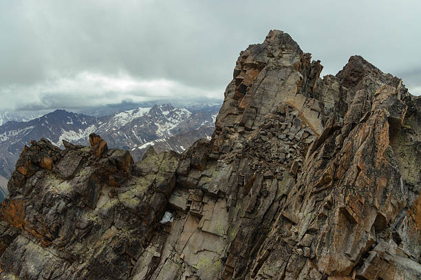 Hikers explore a rocky mountain landscape with steep slopes and sharp peaks. Clouds cover parts of the sky as snow-capped mountains stretch into the distance.