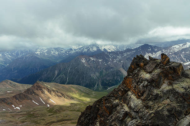 Clouds cover the sky while mountain ranges stretch far into the distance. rocky foreground stands out against the green hills and snow-capped peaks under gray skies.