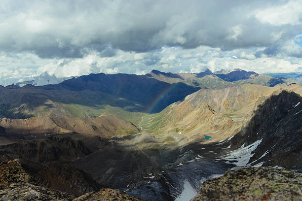 Clouds hover over a vast mountain valley. rainbow arcs across the landscape. Mountains stretch into the distance, with patches of green and snow visible.