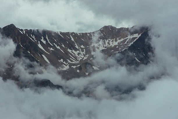 Snow on mountain tops is visible as clouds surround the peaks. scene shows a rugged landscape with rocky formations and low visibility due to fog.