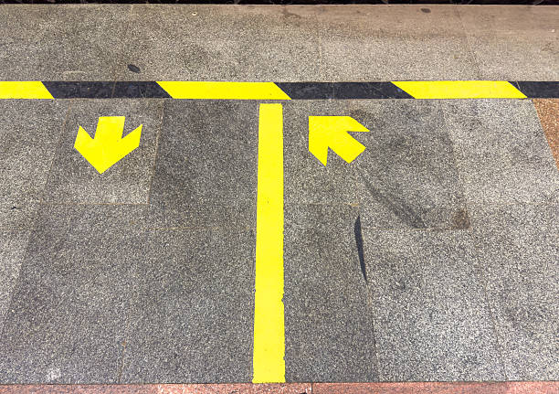 Yellow arrows on a concrete floor guide movement directions in a public area, featuring a textured surface and contrasting colors