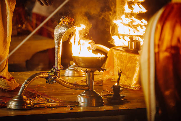 A close-up view of a ceremonial lamp with bright flames during a Hindu fire ceremony in Varanasi, India. The ornate metal vessel and glowing embers create a warm, sacred atmosphere.