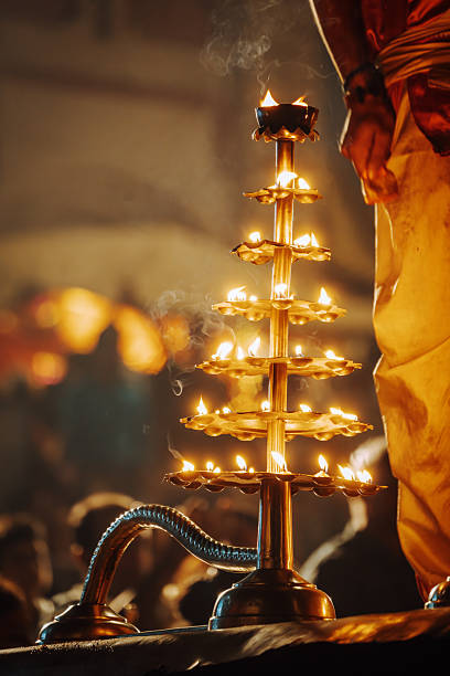 A multi-tiered brass lamp stands alight during a Hindu fire ceremony in Varanasi, casting warm glows. Devotees surround the ritual with reverence and incense smoke rising in the background.