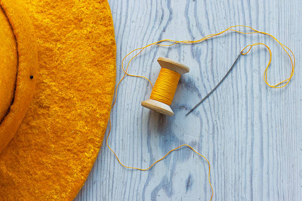 Top view of a yellow thread spool and sewing needle with a loose thread and a felt hat edge on a wooden table