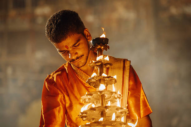 Varanasi, India - November 3, 2024: A devout participant in a vibrant orange robe holds a multi-tier fire offering during a Hindu festival in Varanasi. Warm flames illuminate the scene, conveying devotion, tradition, and spiritual energy.
